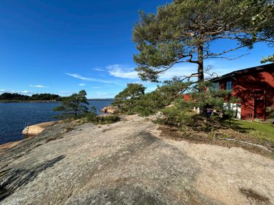 Fotografi av en rød bygning ved vannet med grantrær og en steinete kystlinje.