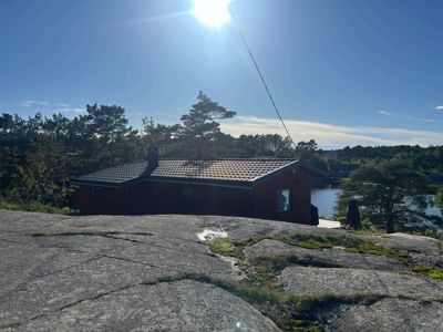 Fotografi av en rød hytte sett fra en høyde, med sollys, trær og vann i bakgrunnen.
