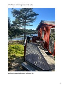 Fotografi av en eksisterende rød hytte med terrasse, sett fra siden. Bildet viser også en del av tomten, en stor gran, vann og nabolag i bakgrunnen.