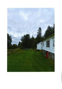 Fotografi av en hvit, langhuset bygning med steinfundament, sett fra siden mot en grønn hage og skog.