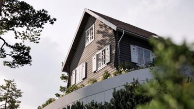 Fotografi av en mørk trehus med hvite vinduer, sett fra en lav vinkel med trær i forgrunnen.