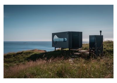 Fotografi av en moderne, mørk hytte eller container med et unikt, asymmetrisk vindu, plassert på en grønn klipperand med utsikt over havet.