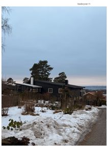 Fotografi av en mørk, moderne enebolig med sne i forgrunnen og et utsikt over vannet i bakgrunnen.