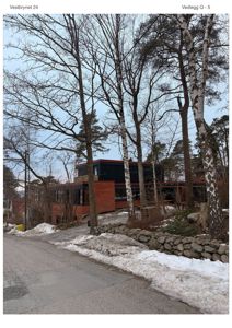 Fotografi av en moderne bygning med rødbrunn fasade og mørke vinduer, sett fra en vei med snø på bakken og trær i forgrunnen.