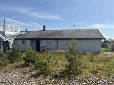 Fotografi av en hvit, langhuset bygning med grå tak, sett fra siden. Bygningen har vinduer, en dør, en skorstein og en flaggstang. Foran ligger en grusete hage med små trær.