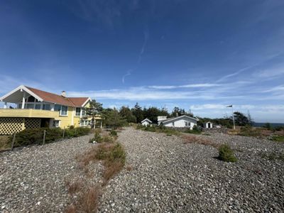 Fotografi av en tomme tomte med grusdekke, omgitt av naboerhus og hav i bakgrunnen.