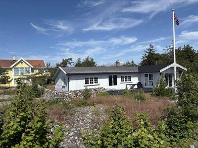 Fotografi av en hvit hytte med grå tak, sett fra siden. I bakgrunnen er det en gul hytte, trær og en blå himmel med skyer. En norsk flaggstang står til høyre.