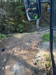 Fotografi tatt fra en kjørende bil (eller lastebil) som viser en grusvei eller terreng i en skogskant med en liten bekke i bakgrunnen.