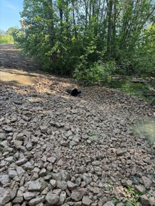 Fotografi av et utgravet område med grus, en svart rørledning og skog i bakgrunnen.