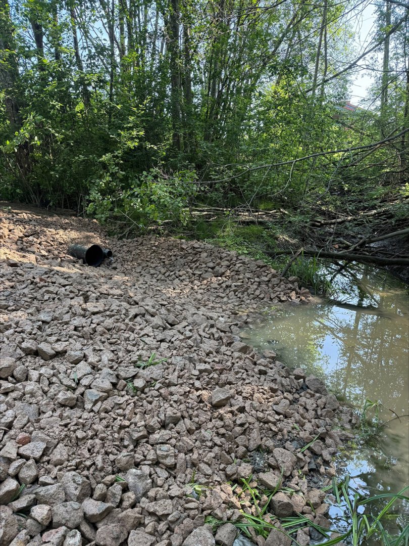 Fotografi av et utendørs område med grus, vann og skog.