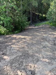 Fotografi av en utgravet eller forberedt tomt med jord og skog i bakgrunnen.