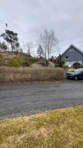 Fotografi av en eksisterende eller planlagt tomt med en grå hus i bakgrunnen, en bil, og vegetasjon.