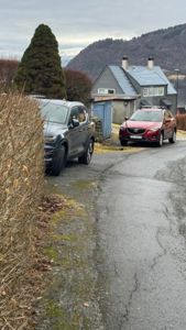 Fotografi av en vei med to biler parkert foran en grå hytte med blå dører og et blått skur. Bakgrunnen viser fjell og trær.