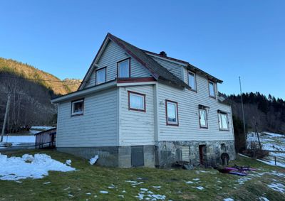 Fotografi av en hvit, toetasje hytte med steinmur i kjelleretasje, sett fra en vinkel som viser både fasade og sidevegg. Bakgrunnen består av snødekket terreng og skog.