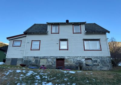 Fotografi av en eksisterende hvit malte hytte med steinmur i kjelleretasje, sett fra front.