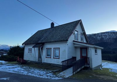 Fotografi av en hvit, trepanelert hytte med skifer-tak sett fra siden. Bildet viser også en rulletrapp til inngangen, vinduer, en skorstein og omgivelsene med snø og fjell i bakgrunnen.