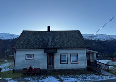 Fotografi av en hvit, enetasjes hytte med rødt vindusrammer, sett fra front. Bildet viser også omgivelsene med snødekte fjell i bakgrunnen.