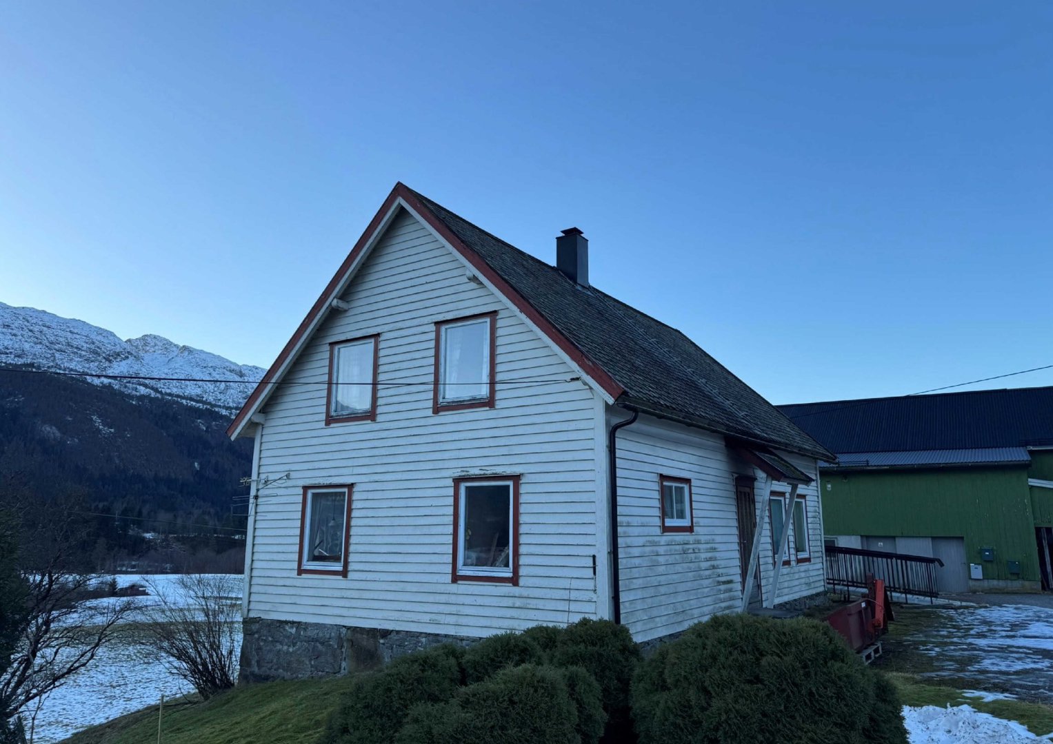 Fotografi av en hvit, toetasjes hytte med steinmur i bunnen, sett fra siden. Bakgrunnen viser snødekte fjell og en annen bygning.