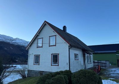 Fotografi av en hvit, toetasjes hytte med steinmur i bunnen, sett fra siden. Bakgrunnen viser snødekte fjell og en annen bygning.