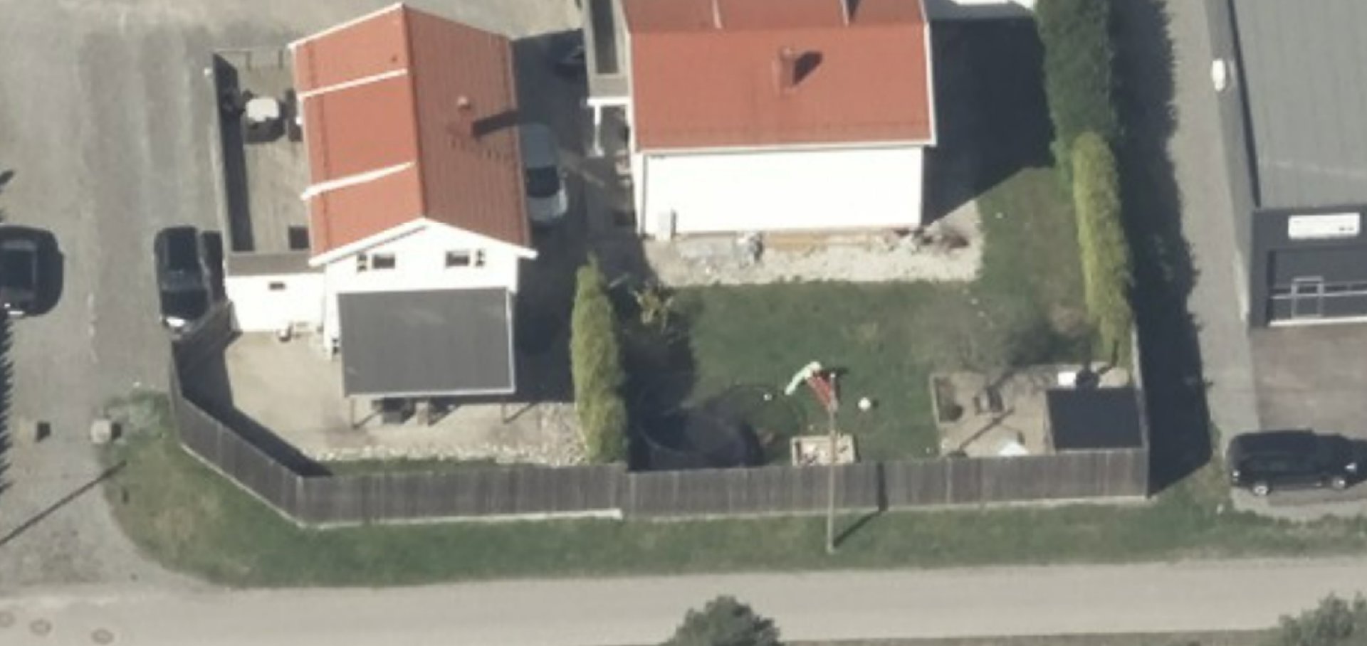 Luftfoto (aerial view) av en tomt med to hus, en hekk og parkerte biler.