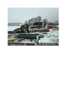 Fotografi av et byggeplass under arbeid i snø. Bildet viser en grøft, en graver, byggematerialer (bl.a. isolasjon), og en delvis ferdig bygning i bakgrunnen.
