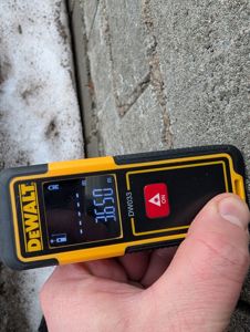 Fotografi av en person som holder en laseravstandsmåler (DeWalt DW033) opp mot en betongvegg. Måleren viser en avstand på 3,650 meter.