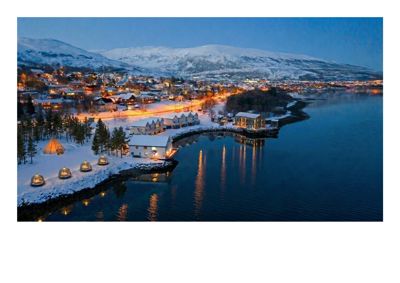 Fotografi av en bygning ved vannet med snødekte fjell i bakgrunnen.