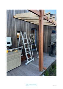 Fotografi av en utvendig terrasse eller veranda under bygging, med en arbeidstaker som monterer en trekonstruksjon.