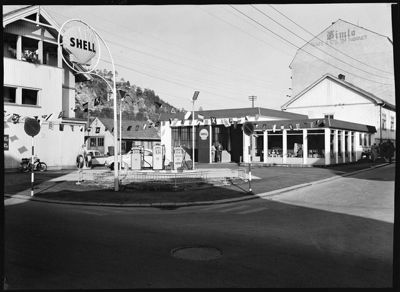 Sort-hvitt fotografi av en bensinstasjon med et SHELL-skilt og en bygning i bakgrunnen.