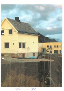 Fotografi av en eksisterende bygning (gult hus) sett fra vest mot sør, med en annen bygning i bakgrunnen.