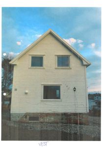 Fotografi av en hvit, toetasjes bygning sett fra vest. Bildet viser fasaden med vinduer, tak og grunnmur, samt omgivelsene med vann og båt i bakgrunnen.