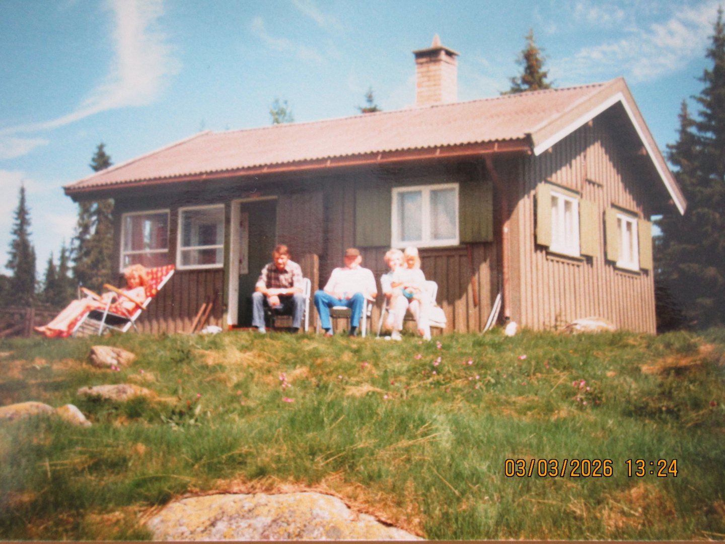 Fotografi av en liten hytte med tre personer sittende utenfor.