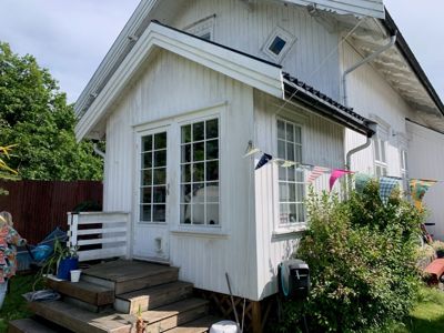 Fotografi av en hvit, trepanelert hytte sett fra siden og foran. Bildet viser fasaden med vinduer, dør, trapp opp til verandaen, takrenner og omgivelser som gress, busker og et hekkverk.