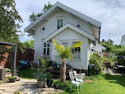 Fotografi av en hvit villa sett fra bakgårdssiden, med hage, møbler og nabolag i bakgrunnen.