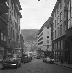 Et svart-hvitt fotografi av en gate i en by, sannsynligvis Bergen, med bilmerker og bygninger fra tidligere århundre.