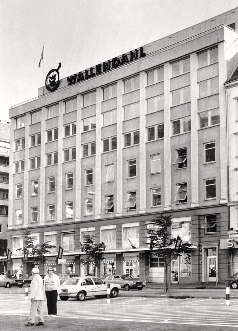 Sort-hvitt fotografi av et fleretasjes bygg med 'WALLEND AHL' skiltet på taket, sett fra gateneiv.
