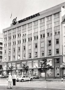 Sort-hvitt fotografi av et fleretasjes bygg med 'WALLEND AHL' skiltet på taket, sett fra gateneiv.