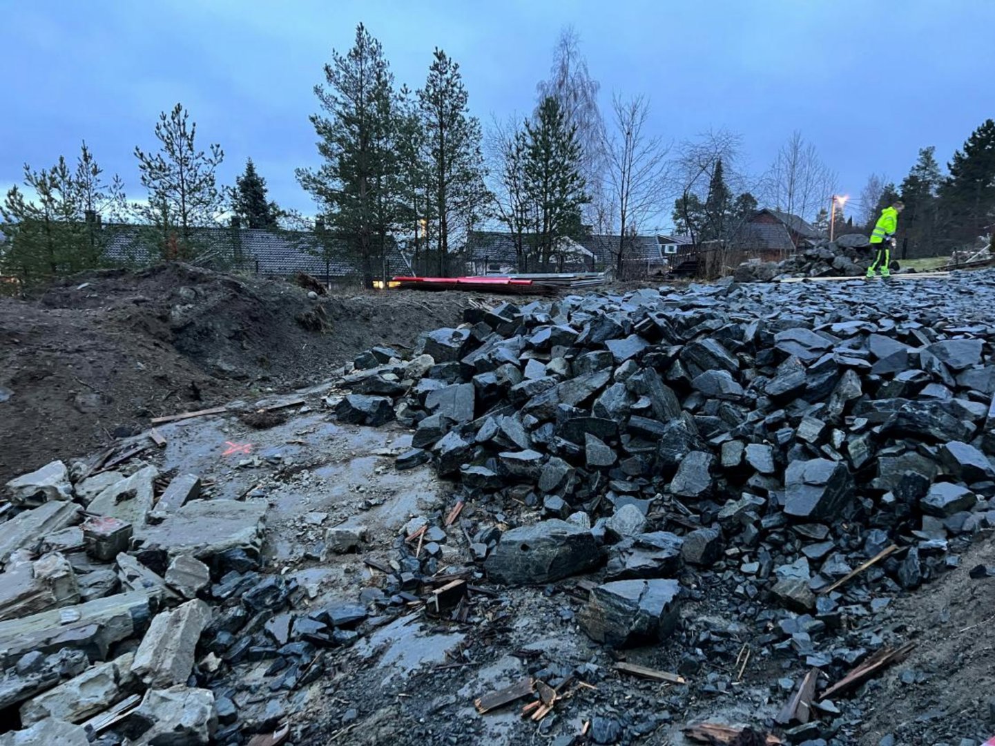 Fotografi av et byggeplassområde med store steinblokker i forgrunnen, en person i sikkerhetsutstyr i bakgrunnen og bebyggelse i distansen.
