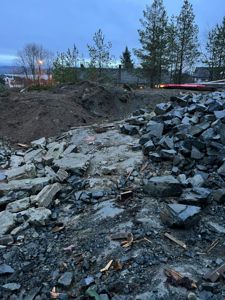 Fotografi av et byggeplassområde med steinhuggeri, grus og en bakgrunn med trær og hus.