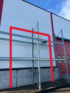 Fotografi av en bygningsside med hvite paneler, en betongfundament, og en metallkonstruksjon (scaffolding) foran. Bildet viser også en del av en rød fasade i bakgrunnen.