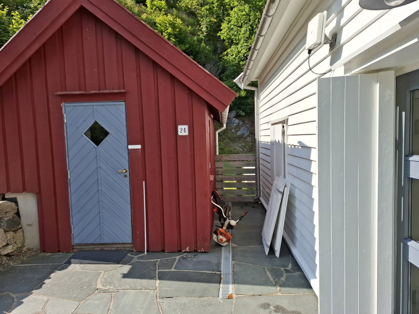 Fotografi av en rød skur med blå dør og en hvit bygning ved siden av, sett fra bakkenivå.