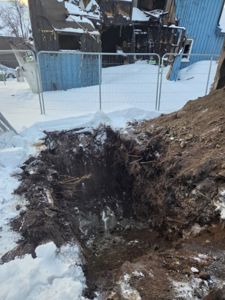 Fotografi av en byggeplass eller skadested med en grøft i forgrunnen og en delvis ødelagt bygning i bakgrunnen.