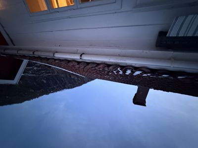 Fotografi av en bygning sett fra nede, som viser takflaten med takteglene, en røykledning, nedløp og fasaden mot en bakke.