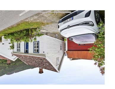 Fotografi av en hvit, trepanelt bygning med blå vindusrammer og et rødt tak, sett fra siden. En hvit bil er parkert foran bygningen.