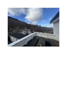 Fotografi av en terrasse med hvit balustrade, ved siden av en hvit bygning, med utsikt over et fjellområde med snø og trær.