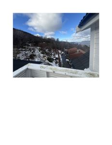 Fotografi tatt fra en balkong eller terrasse som viser utsikt over en snødekt skråning, nabolag med tak og fjord i bakgrunnen.