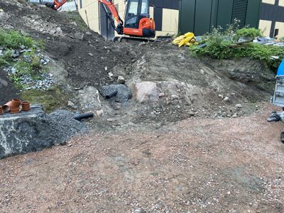 Fotografi av en byggeplass med utgravd terreng, en graver og byggematerialer.