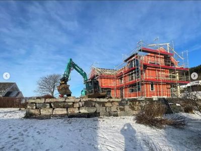 Fotografi av en bygning under oppføring med rødt treverk, stålkonstruksjon og utvendig stålrekkverk. En grønn gravemaskin er synlig ved siden av bygningen, og det er snø på bakken.