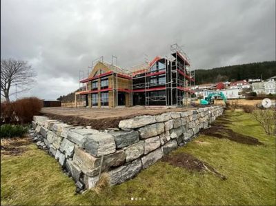 Fotografi av en bygning under oppføring med stålramme, fasadebelegg og utvendig trapp, sett fra et lavt nivå mot en steinmur.