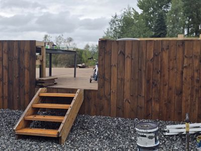 Fotografi av en uteområde under eller etter montering av en terrasse og hekk. Bildet viser en trapp opp til en dekket terrasse, en hekk i forgrunnen og en traktor i bakgrunnen.